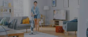 Young Beautiful Woman in Jeans Shirt and Shorts is Vacuum Cleaning a Carpet in a Bright Cozy Room at Home