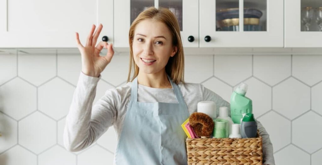 Housemaid from cleaning services holding a box with detergents and household chemical products, showing ok sign, standing on kitchen